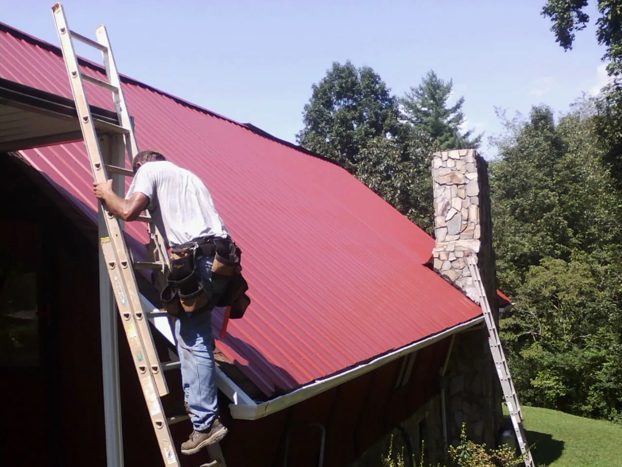 Roofer on a ladder beside a red metal roof and stone chimney — Soffit & Fascia Repair by Roofing Brothers in Corbin